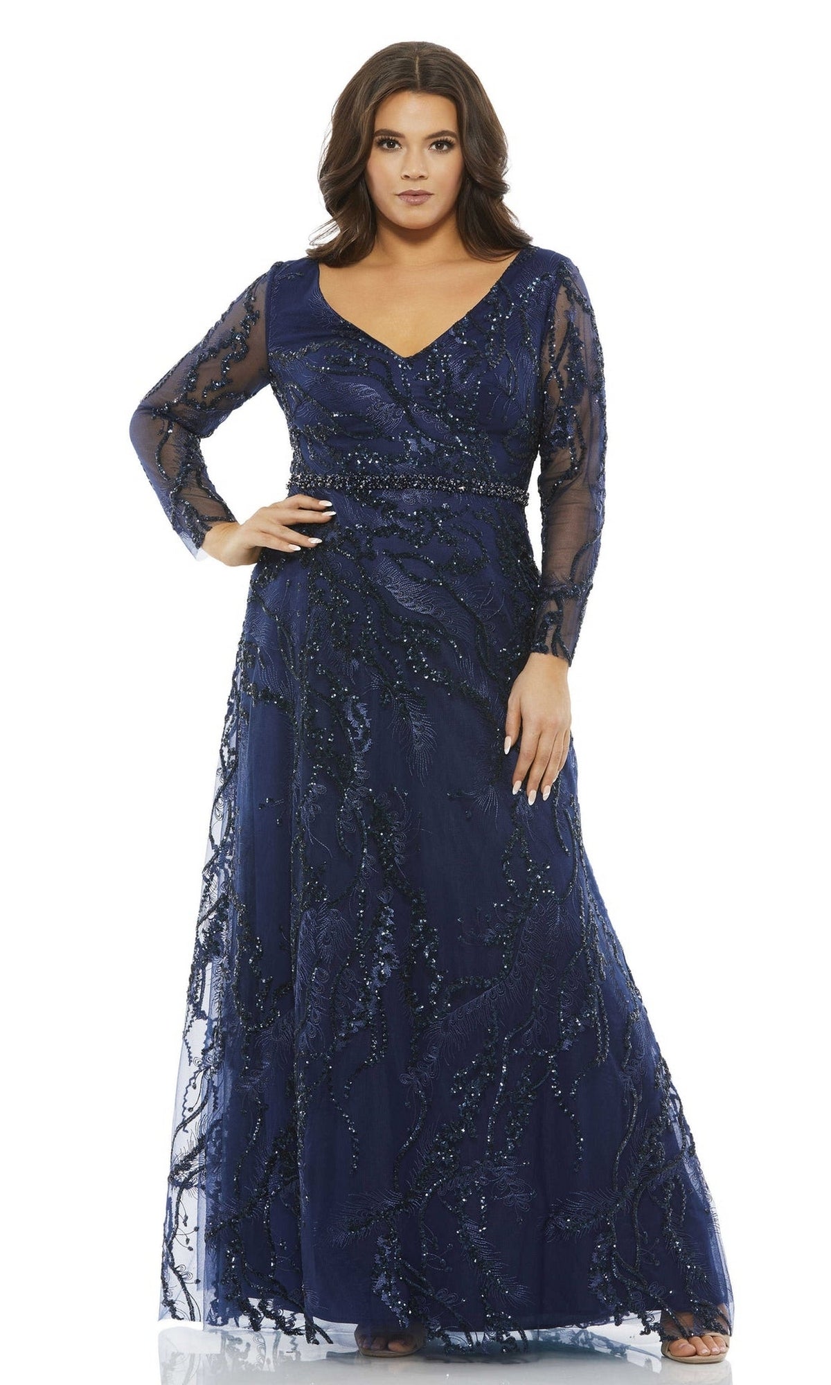 A woman stands against a white background wearing the Long Plus-Size Prom Dress 67922 by Mac Duggal, a navy blue, floor-length A-line gown with long sleeves and sequin embellishments.